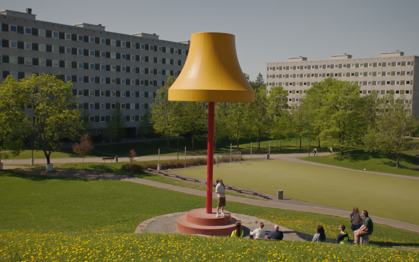 Stills fra film. En stor lampeskulptur i et grønt landskap. Rundt den sitter en gruppe mennesker.