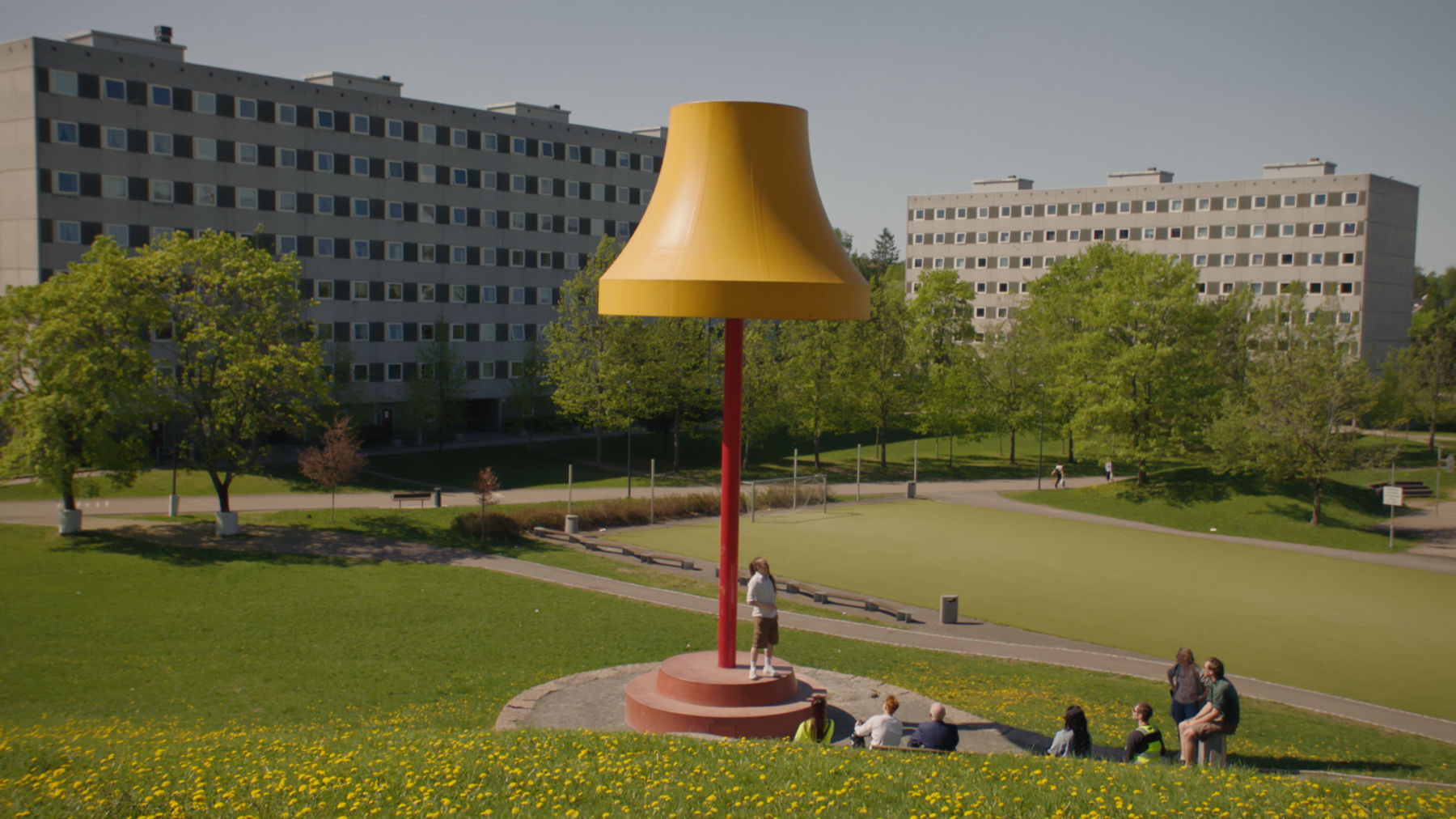 Stills fra film. En stor lampeskulptur i et grønt landskap. Rundt den sitter en gruppe mennesker.