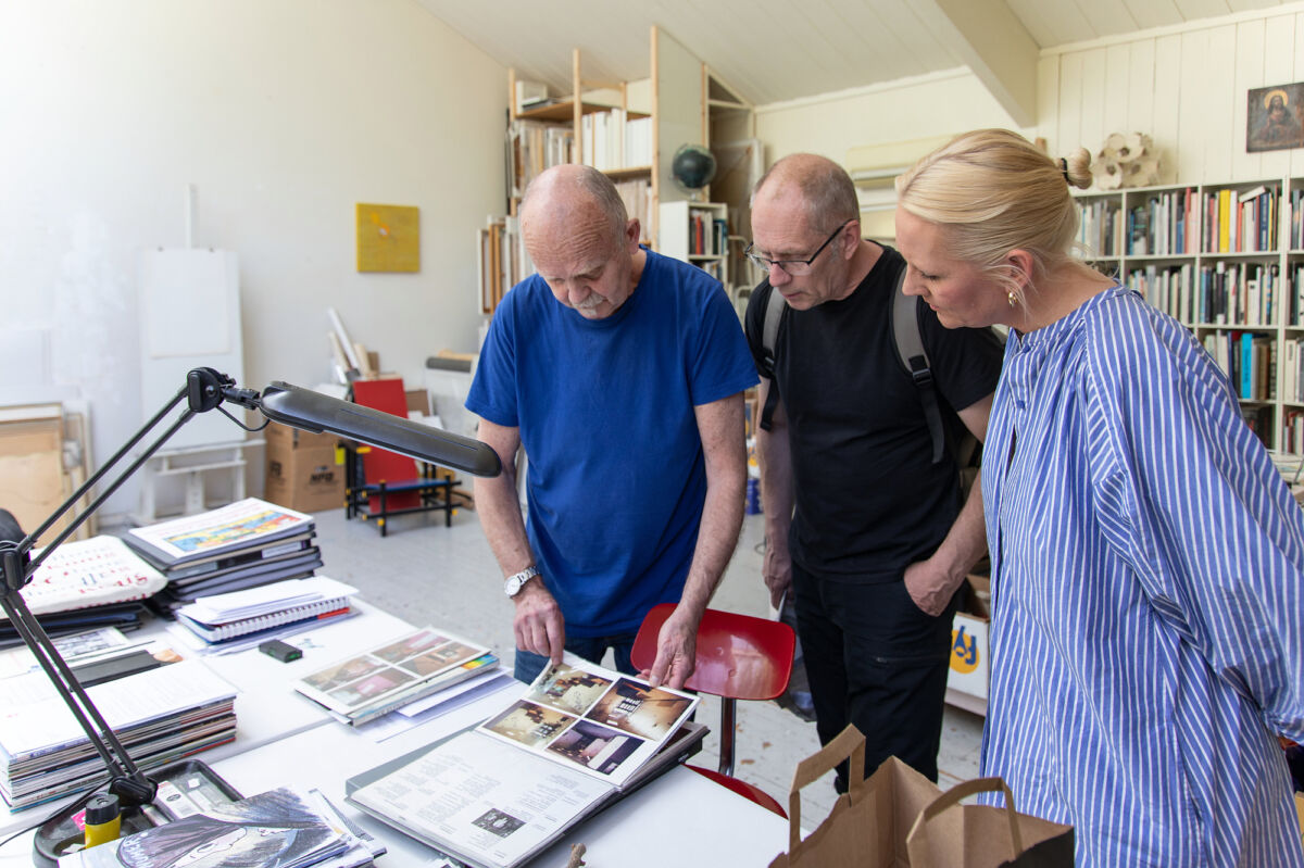 Foto av tre personer som står i et studio og studerer papirer liggende på et bord.