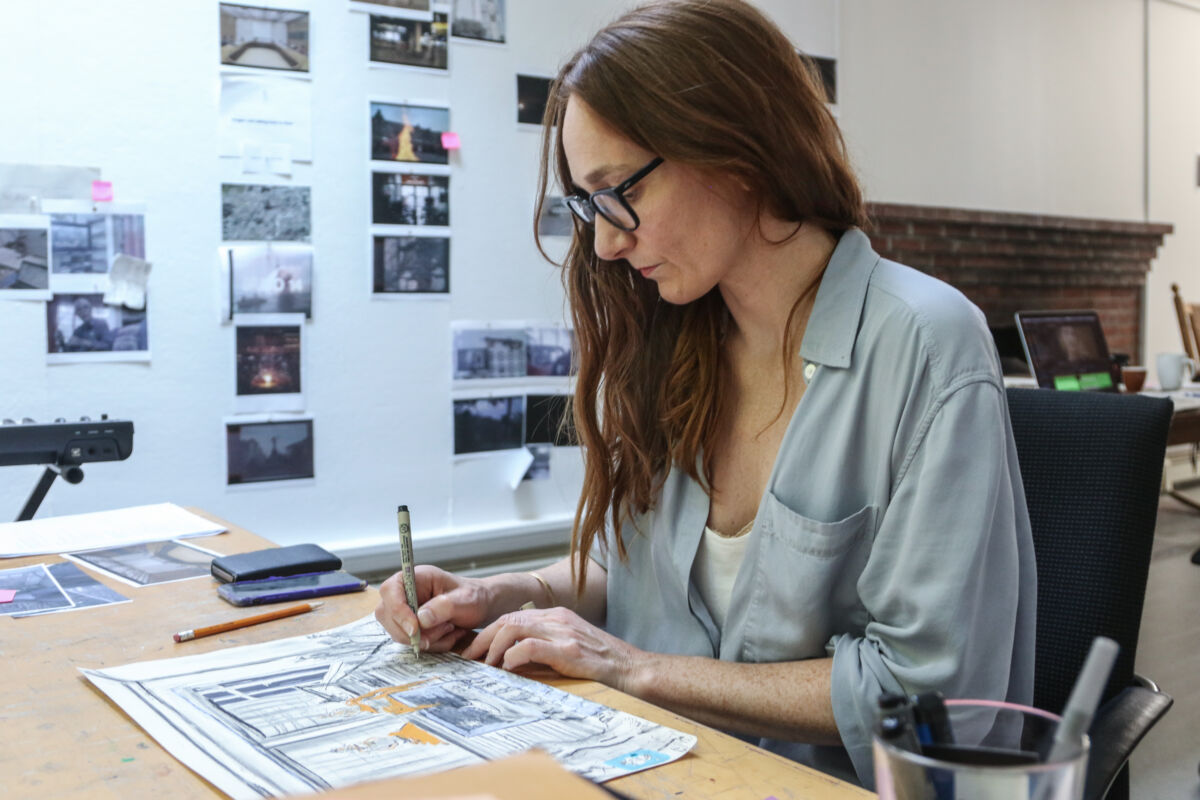 Foto av person som sitter og tegner ved et arbeidsbord.