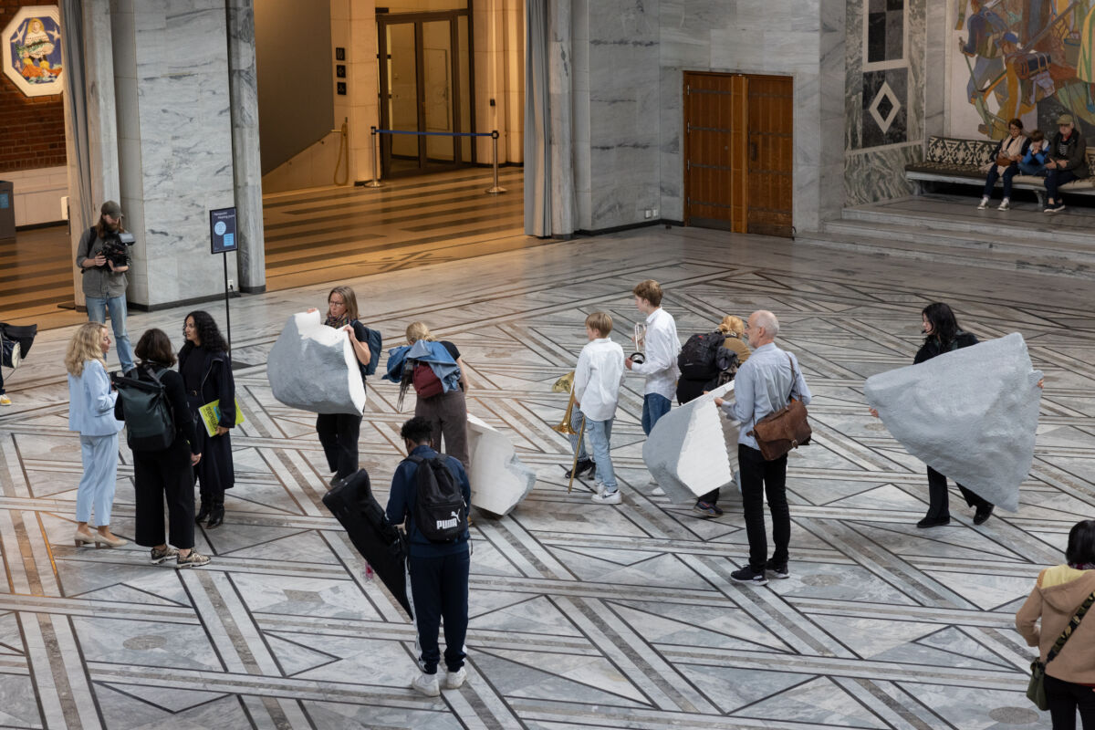 Mange personer stående inne i Oslo rådhus mens de bærer på steinskulptur.