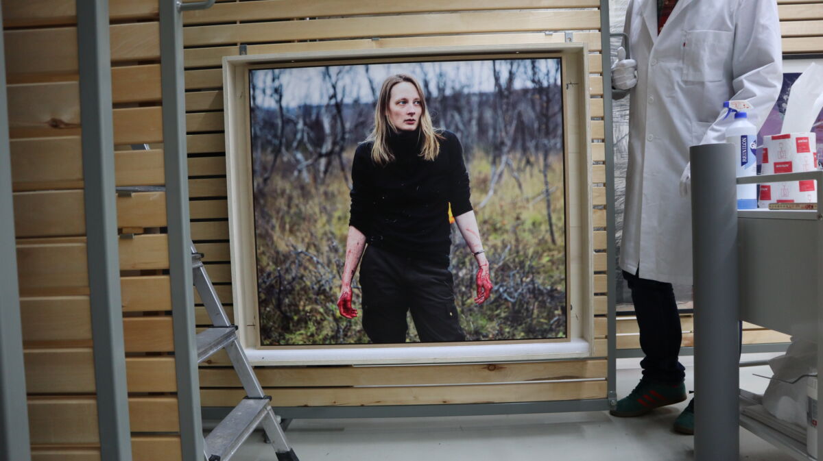 Foto av et kunstfotografi hengende på en skjermvegg i et magasin. En person i hvit frakk står ved siden av.