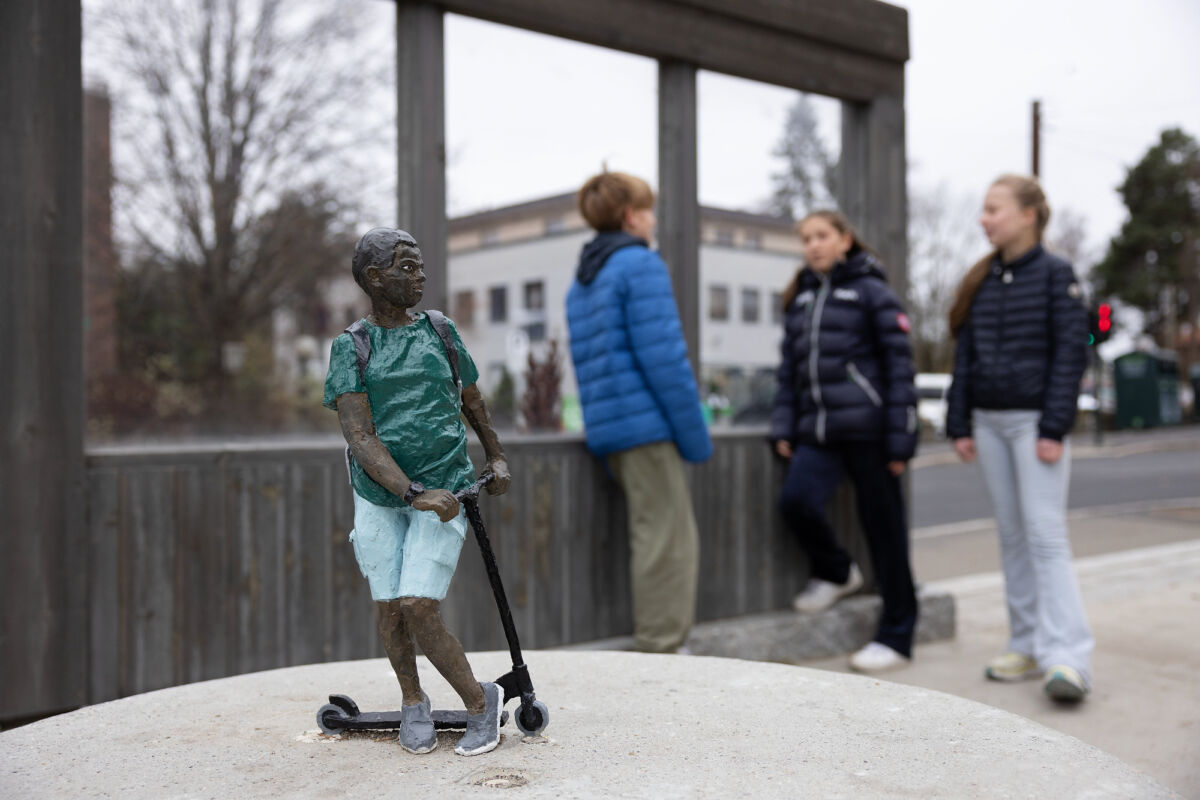 Foto av en skulptur av en ungdom på sparkesykkel. I bakgrunnen står tre elever og snakker sammen.